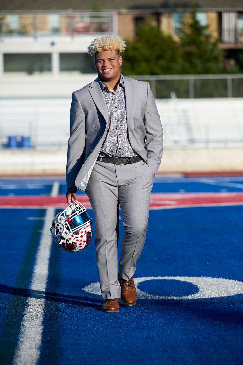 parish episcopal football player senior wearing a gray suit with helmet for senior pictures on the blue football field in dallas by photographer Jeff Dietz